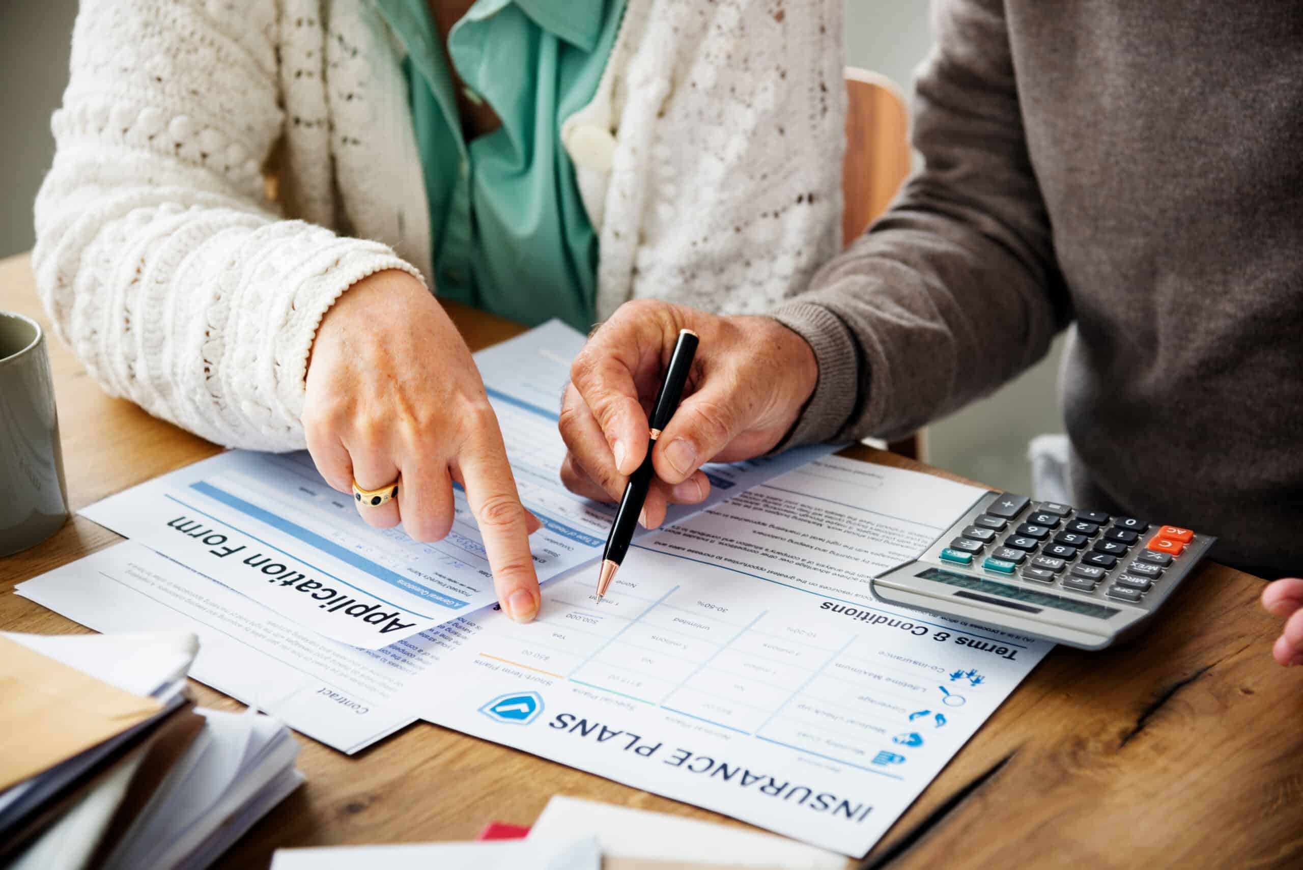 Two people at a table review insurance documents; one points to a form as the other holds a pen. Calculator and papers nearby.