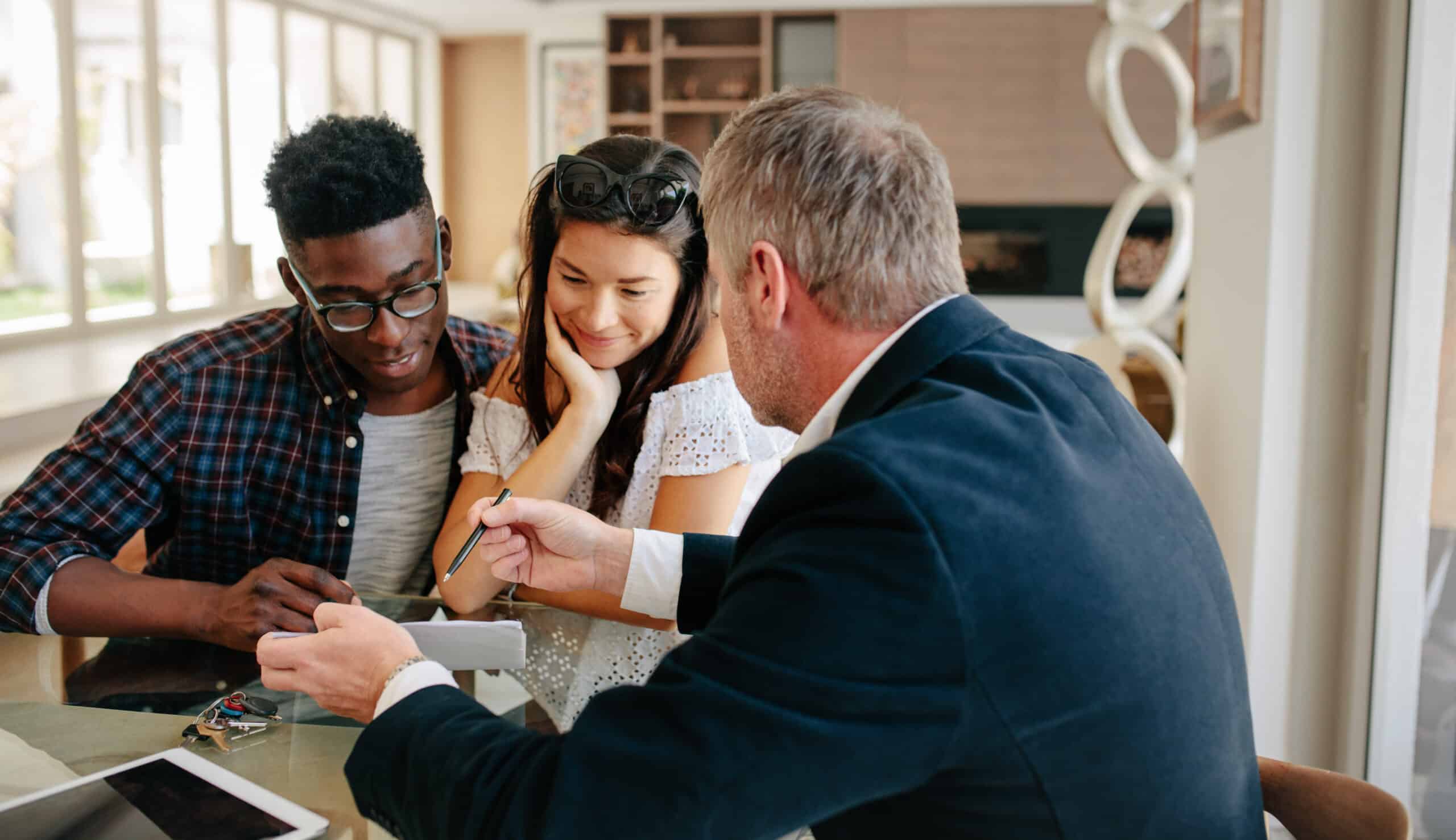A suited man explains paperwork to a young couple at a table in a bright, modern office. They focus intently on the documents.
