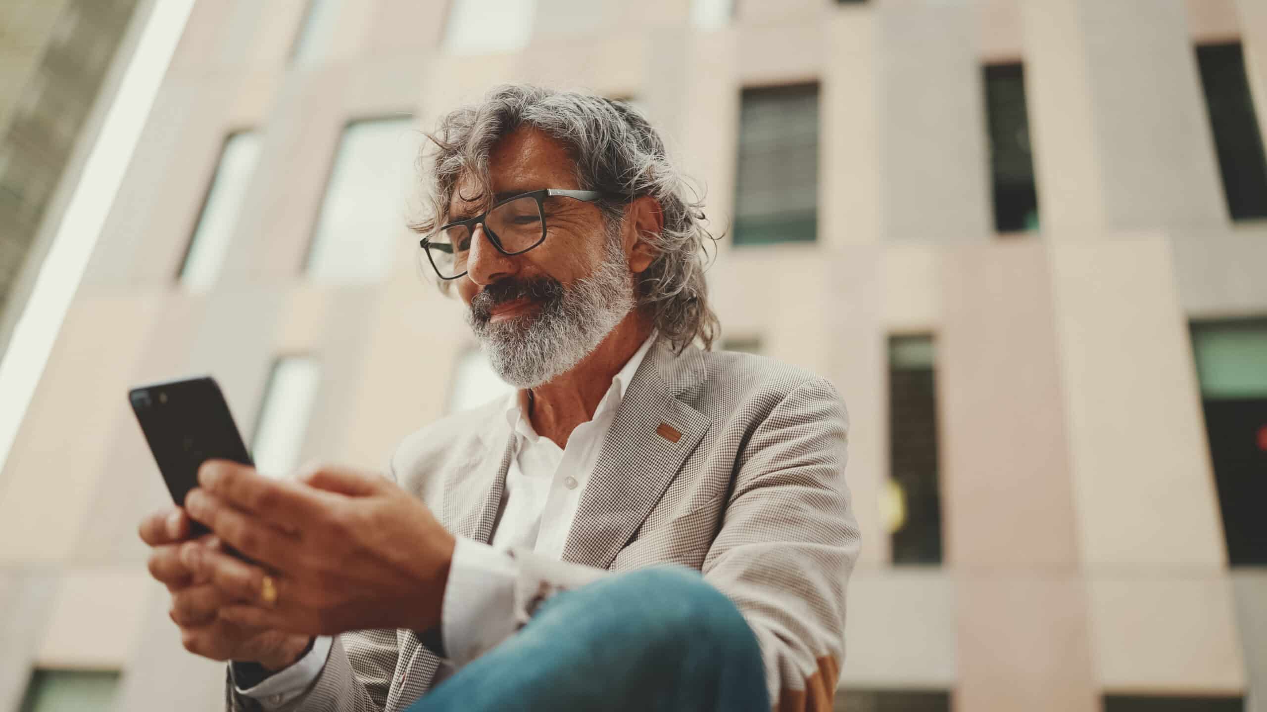 An older man with gray hair and a beard, wearing glasses and a light blazer, sits outside a modern building, smiling at his phone.