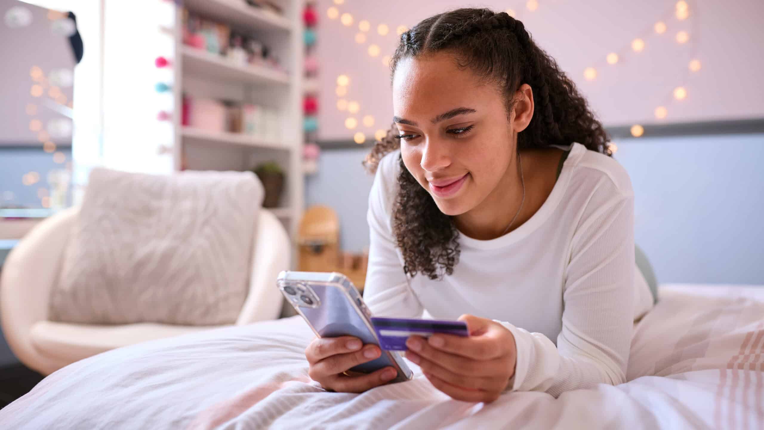 A young woman smiles on her bed, holding a smartphone and credit card, in a softly lit room decorated with string lights.
