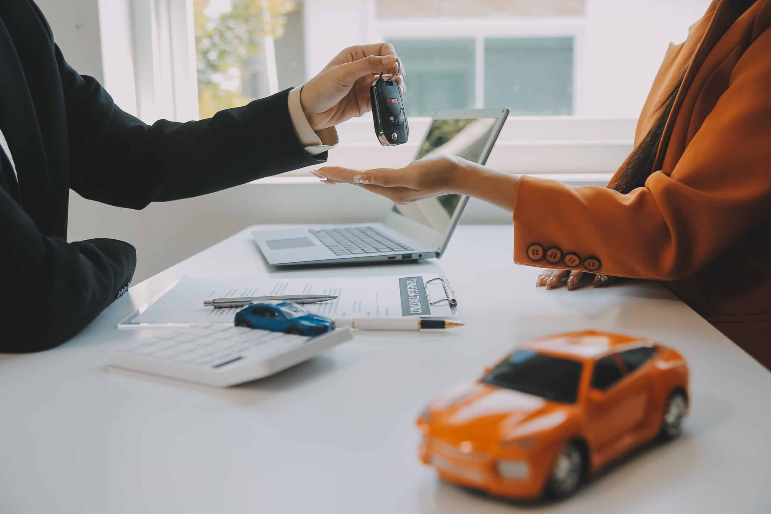 Two people at a desk with a laptop, contract, and toy cars. One hands car keys to the other, suggesting a car sale or lease.
