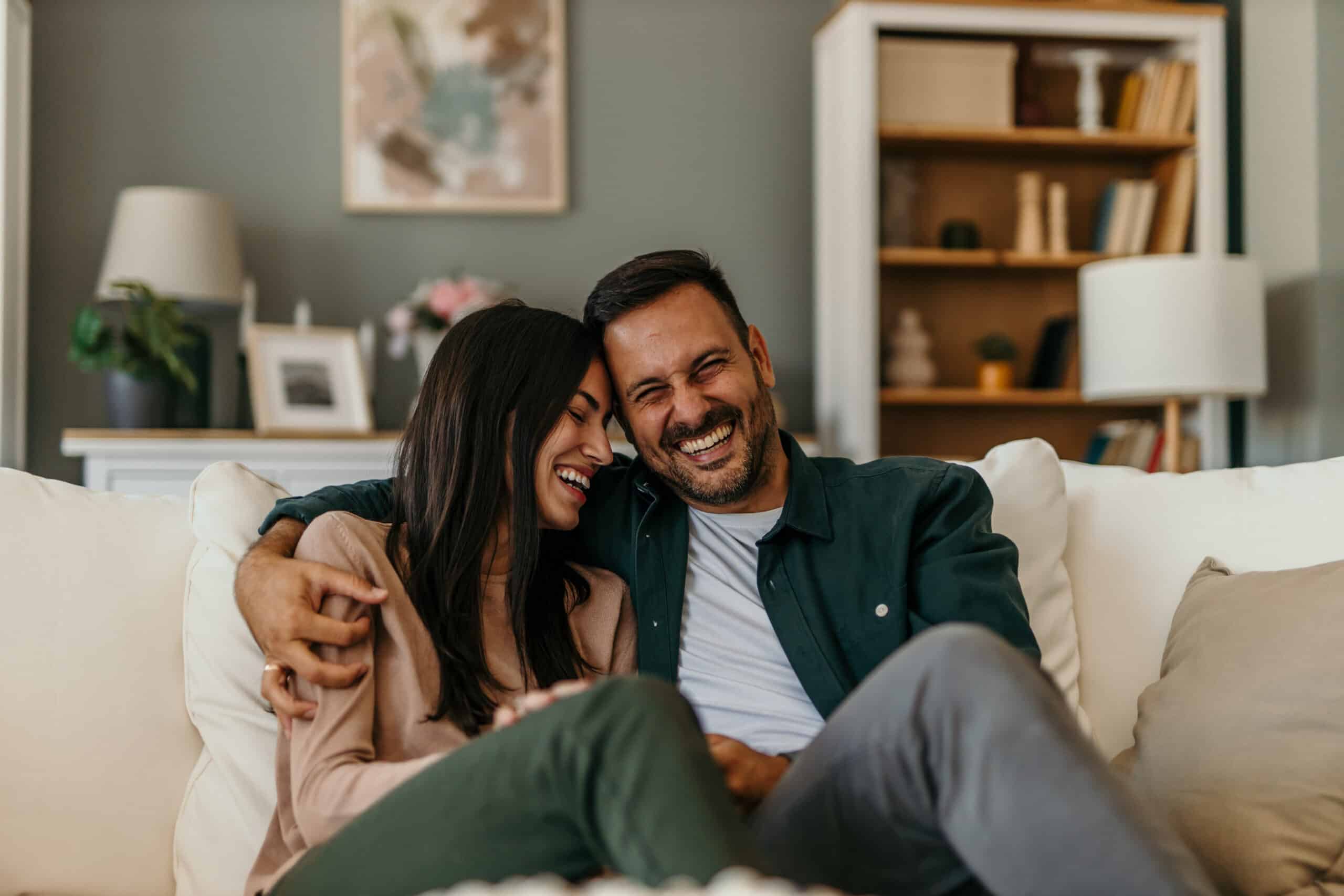 A couple sits together on a couch, smiling. Books, plants, and decor fill shelves behind them, giving the living room a cozy feel.