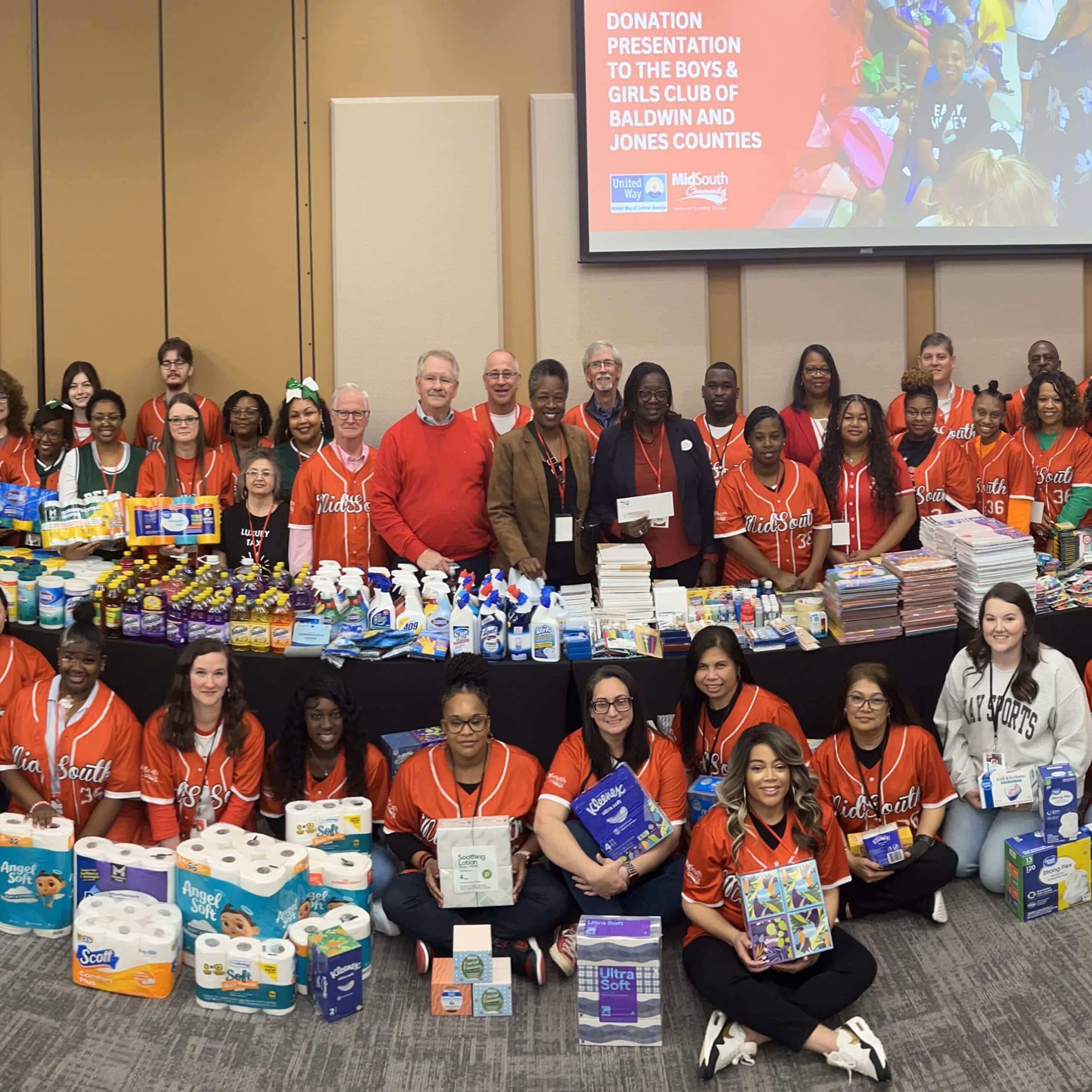 A large group of people, some in red shirts, stand behind tables filled with supplies and a donation sign displayed in the background.