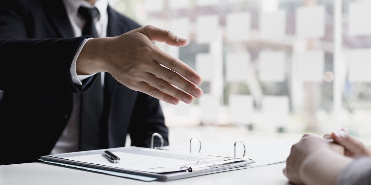 A person in a suit extends a hand for a handshake across a desk with papers and pen; another person's hands rest opposite.