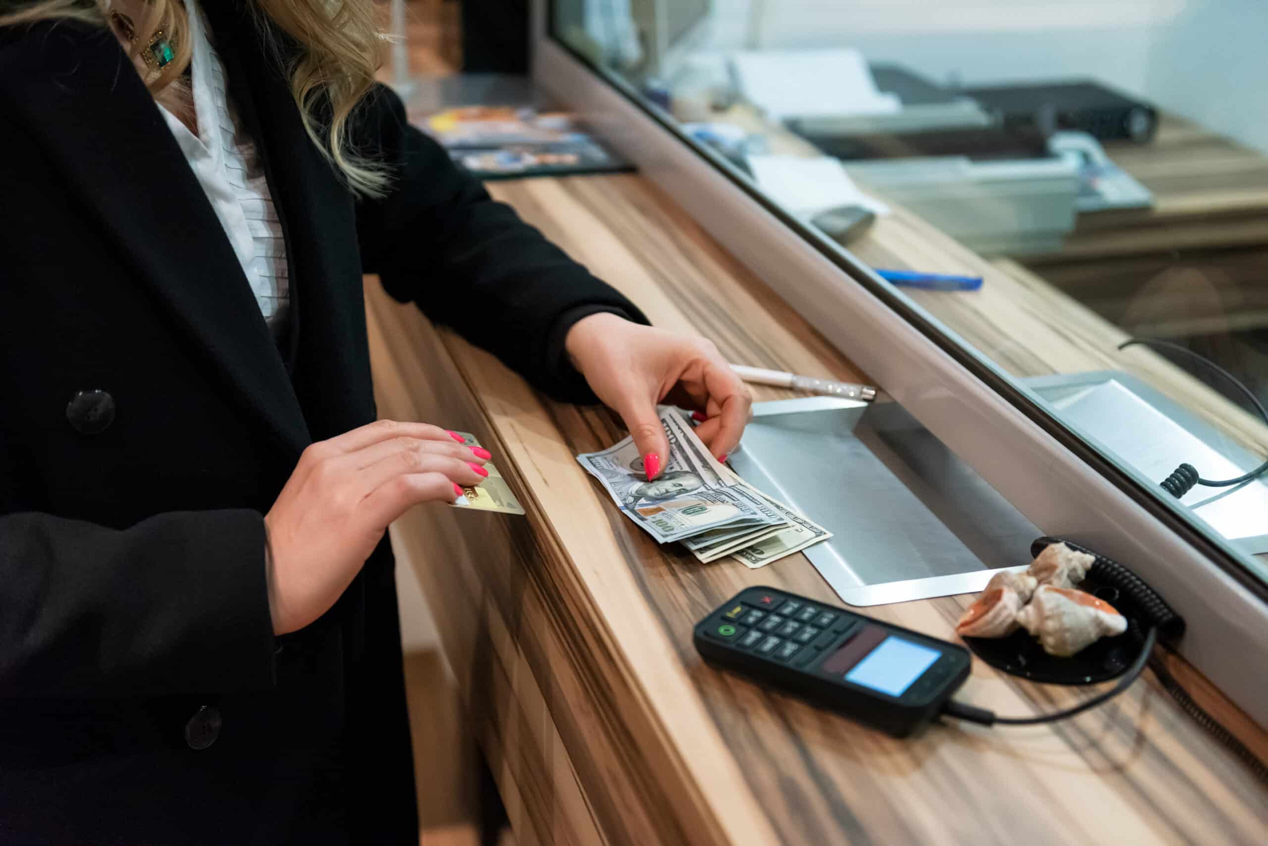 A person at a counter holds a credit card and counts U.S. dollar bills. A card reader, seashells, and papers are on the wooden counter.