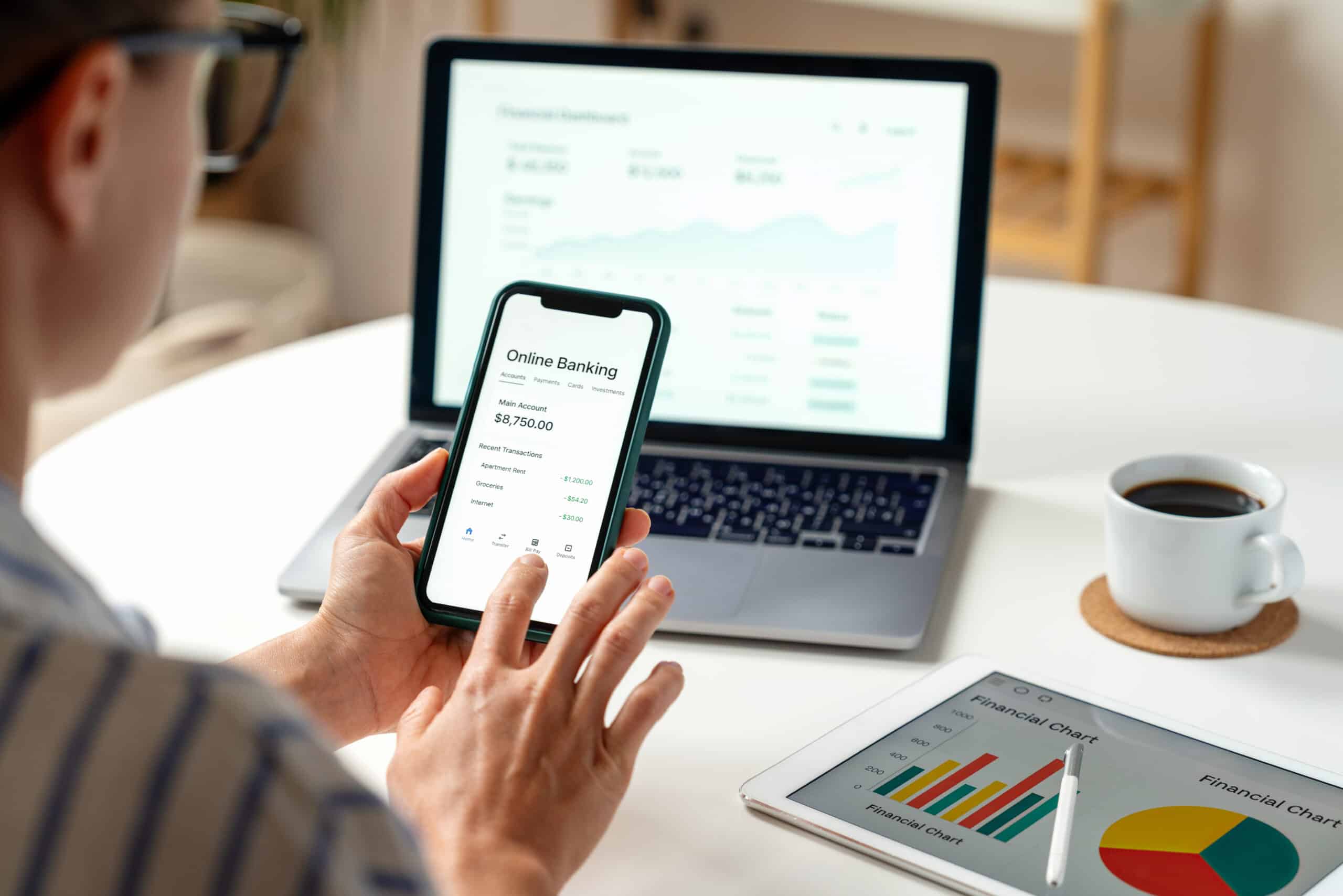 A person uses a smartphone for online banking at a desk with a laptop showing financial data, a tablet with charts, and coffee.
