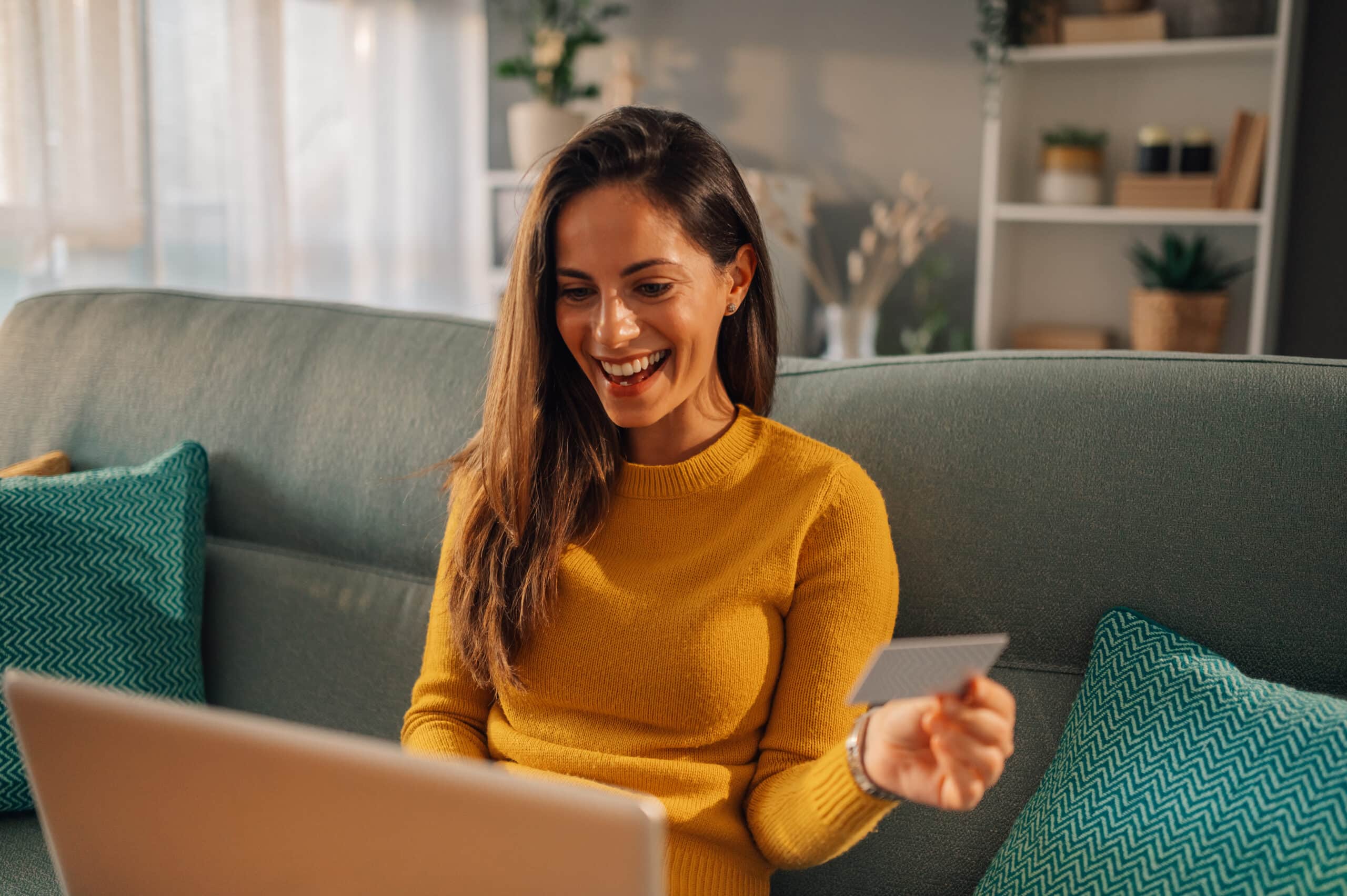 A woman in a yellow sweater sits on a sofa, smiling at her laptop and holding a credit card, shopping online at home.
