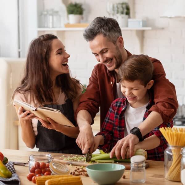 A smiling family of three cooks together in a bright kitchen. Father and son chop veggies as mother holds a cookbook by fresh ingredients.