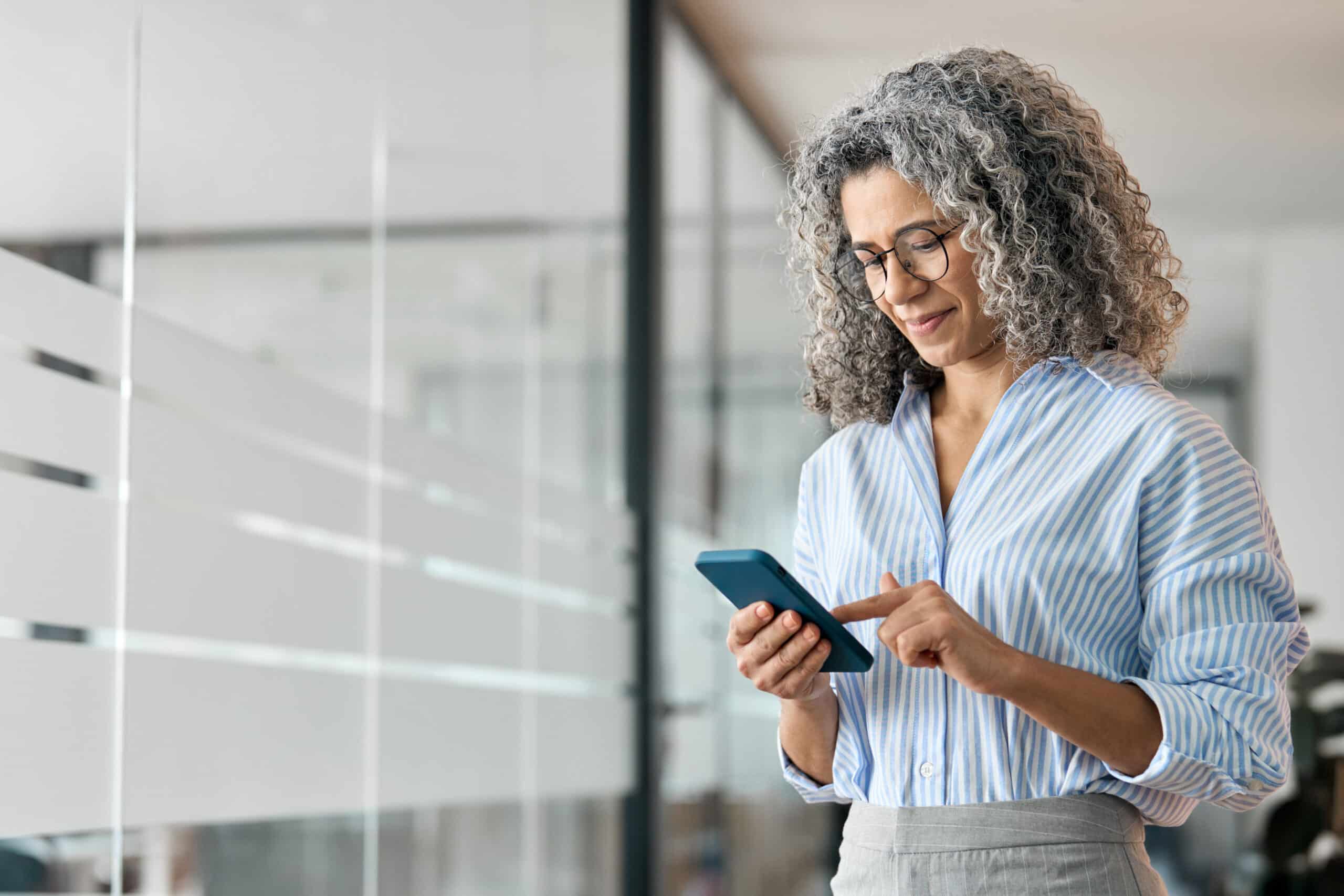 A woman with curly gray hair and glasses stands indoors, smiling at her smartphone in a modern office with a glass wall behind her.