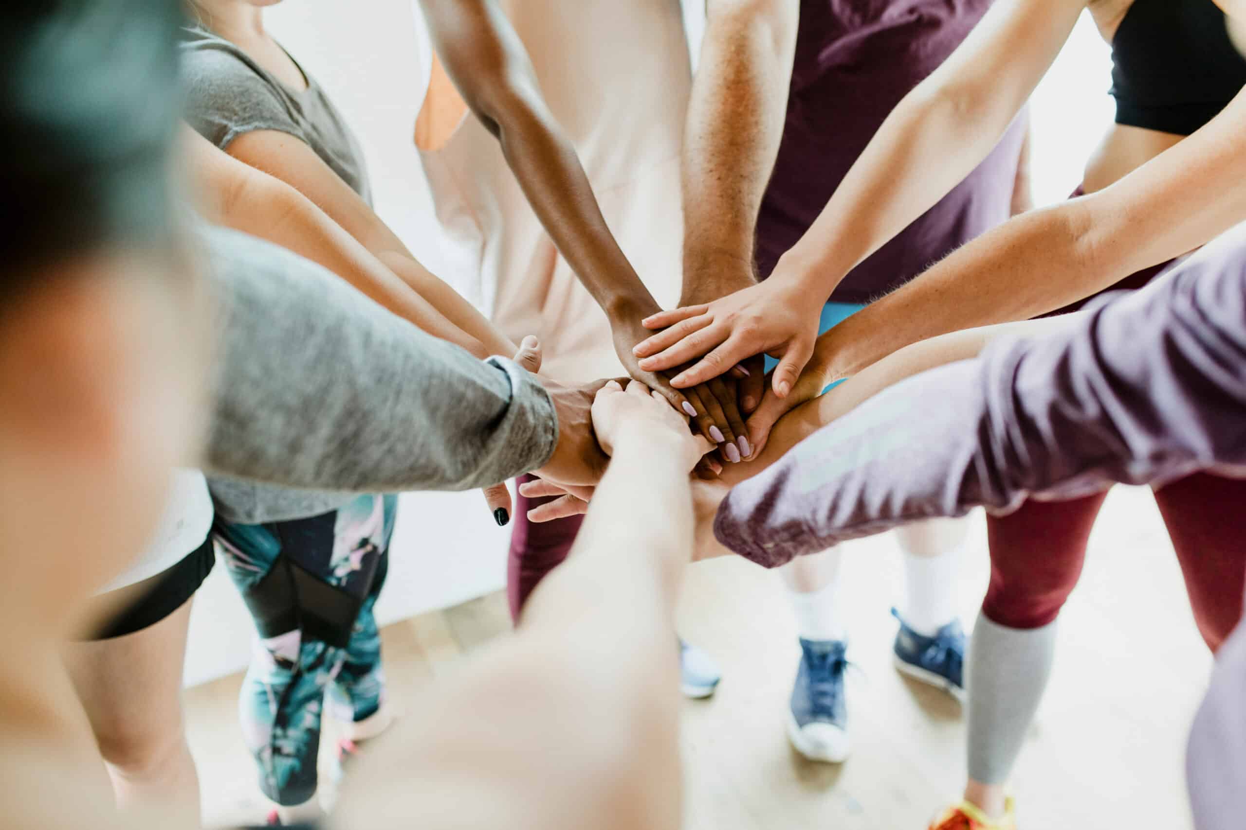 A group of people in a circle extend their arms, stacking hands in the center to show unity. Bright, casual scene, activewear visible.