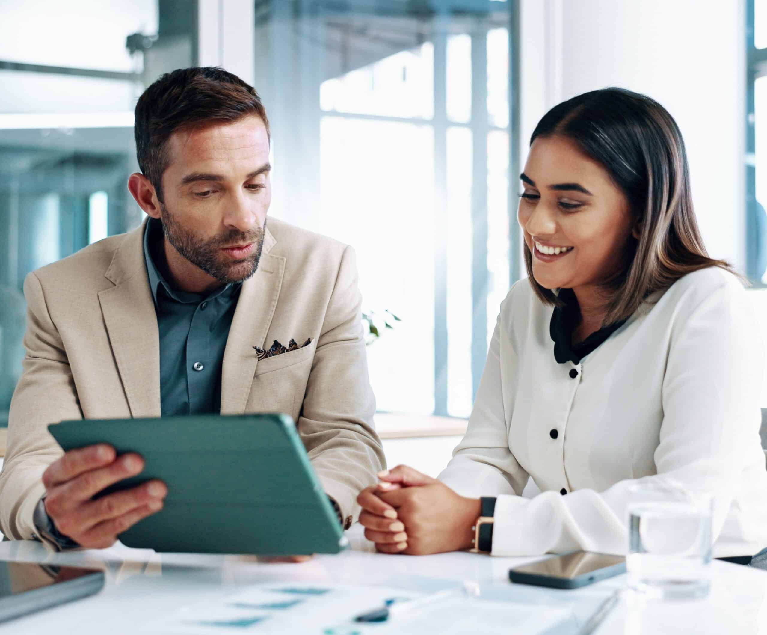 A man in a beige blazer shows info on a tablet to a smiling woman in white at a desk with papers, phone, and glass of water.