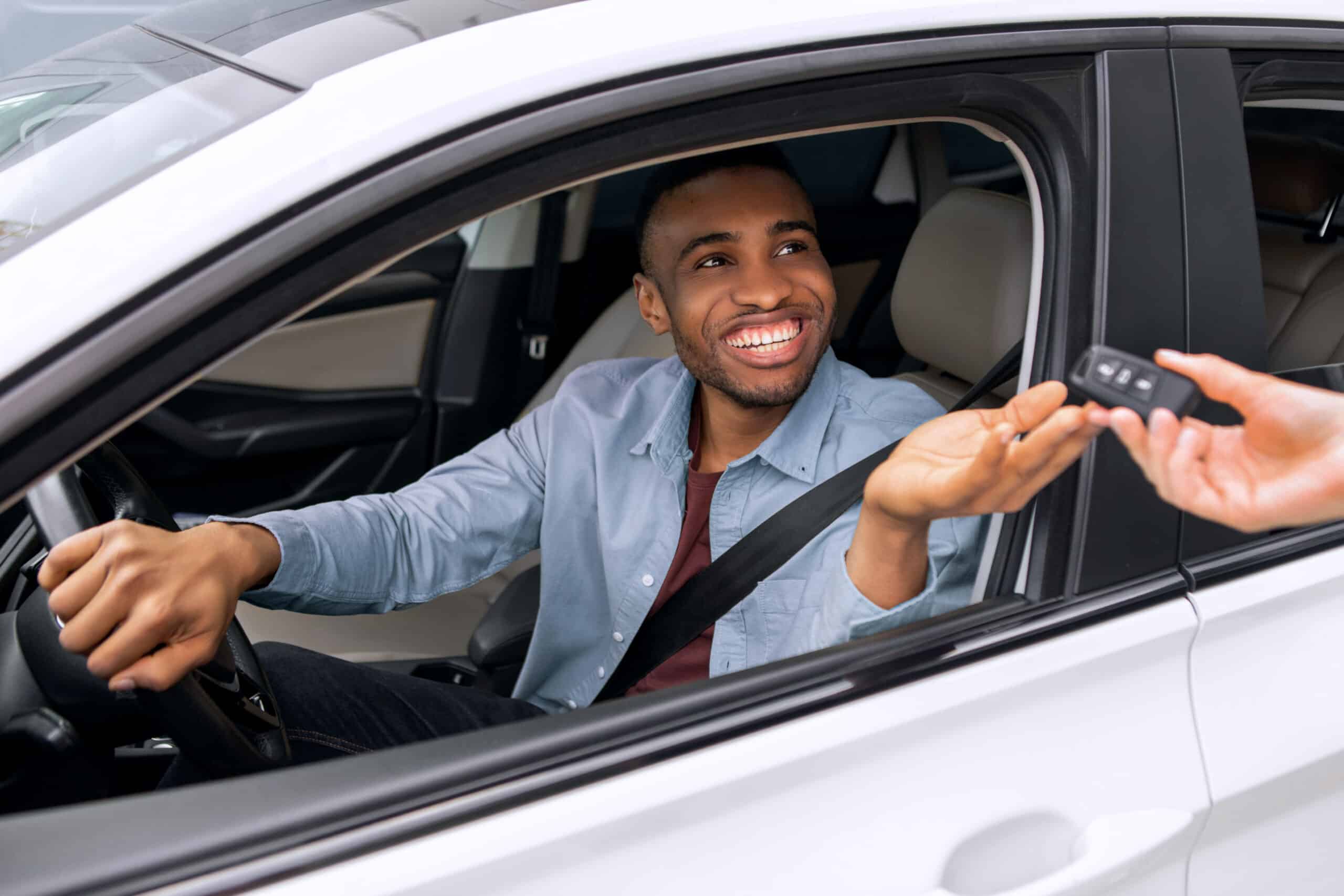 A smiling man in the driver’s seat of a white car reaches out to take a key fob from someone standing outside the vehicle.