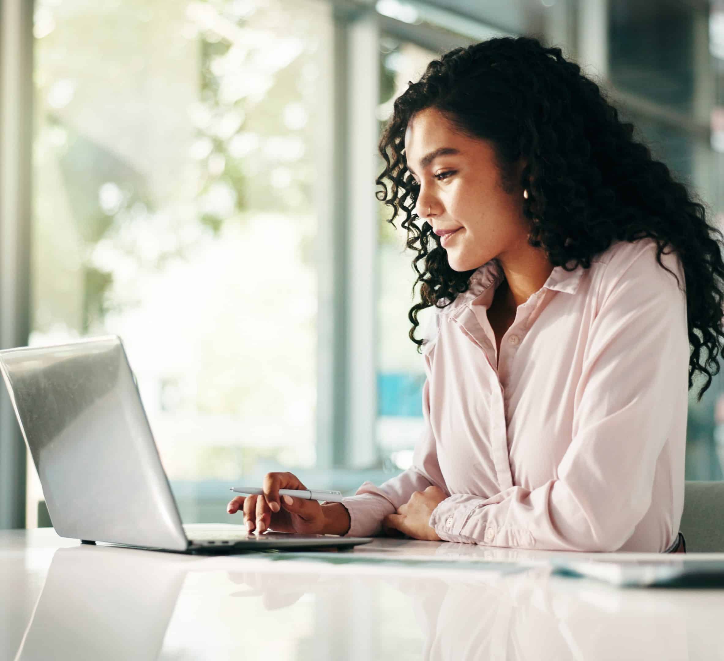 A woman with curly hair smiles at her desk, working on a laptop in a bright office, holding a pen with papers beside her.