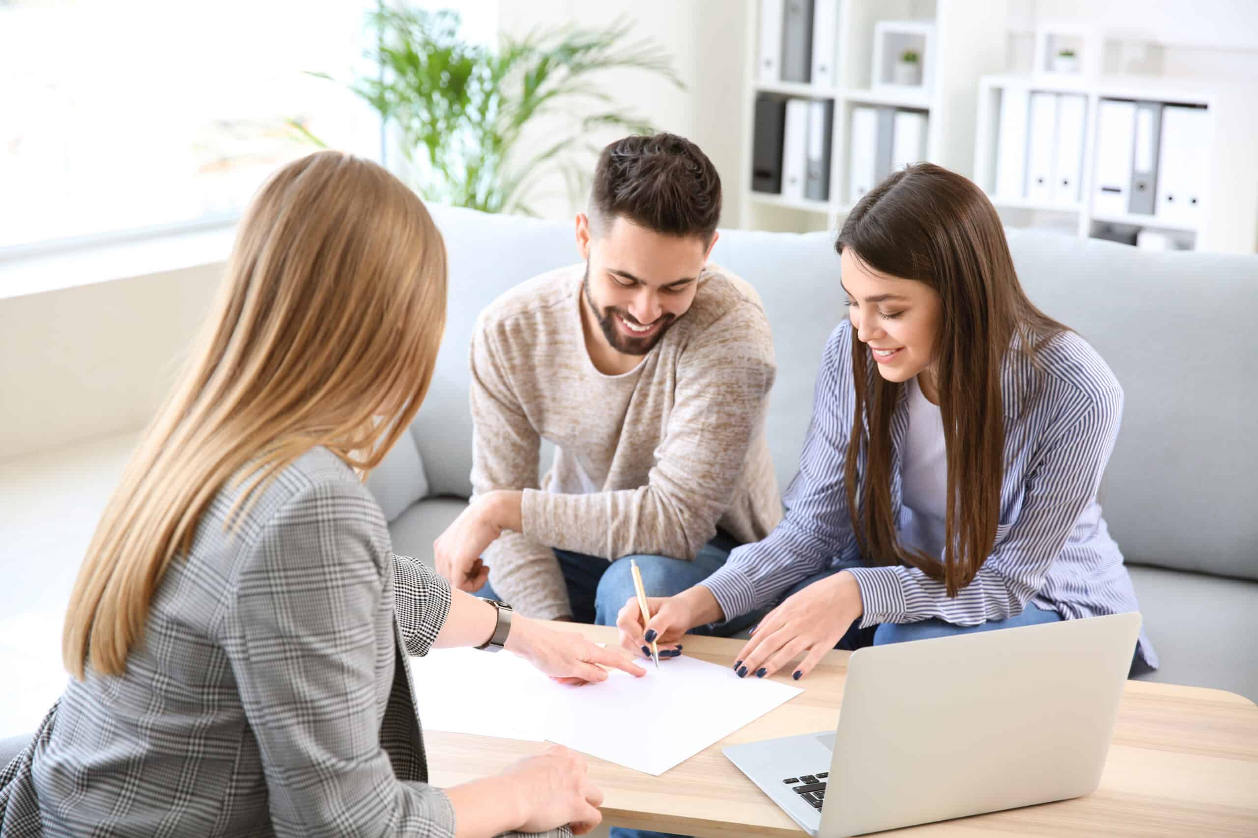 Three people sit around a table discussing paperwork with a laptop open. One woman writes while others smile in a bright office.
