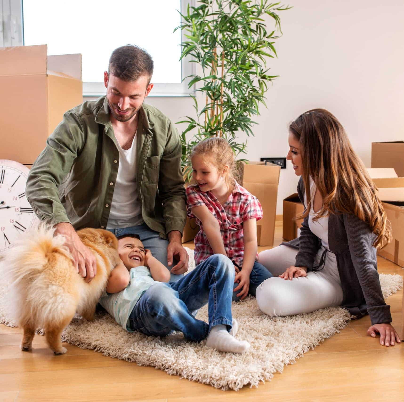 A family of four sits on a rug amid moving boxes, smiling and playing with their fluffy dog in a sunlit room.