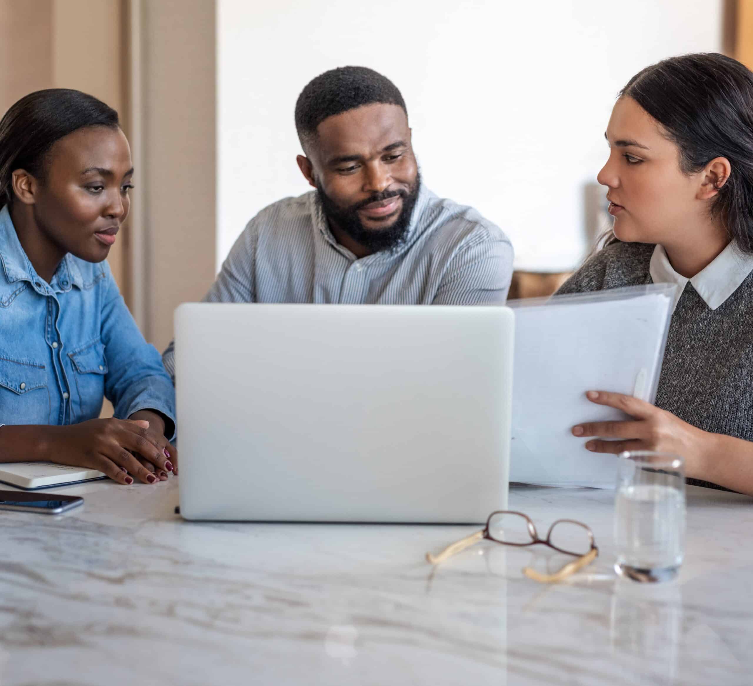 Three people sit at a table with a laptop and documents. One holds papers; others listen. Notebook, glass of water, eyeglasses visible.
