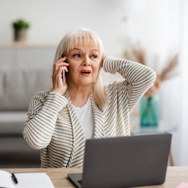An older woman at a desk with a laptop holds a phone, looking worried and touching her head. Papers, plants, sofa, shelves visible.