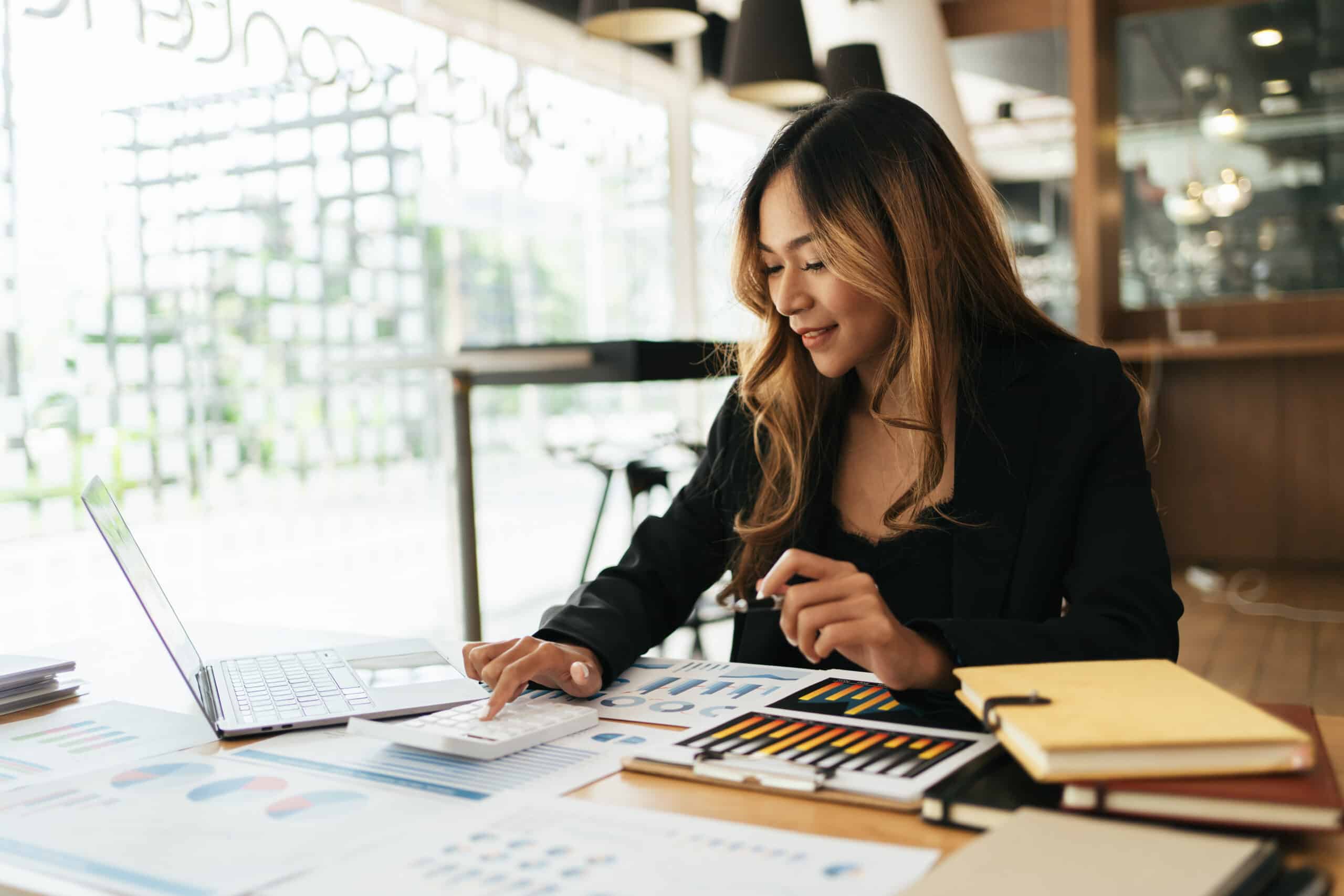 A woman in business attire reviews charts and graphs on a laptop with colorful documents and notebooks spread on her well-lit desk.