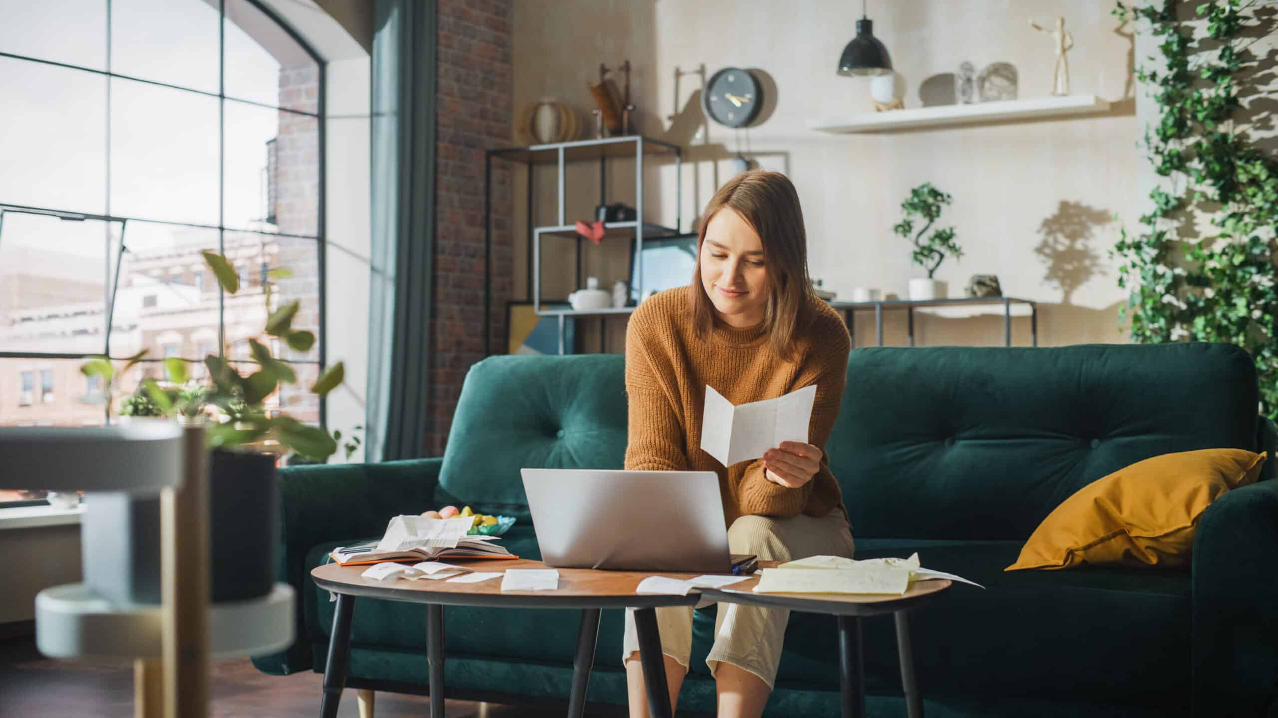 A woman on a dark green sofa reads a paper and uses a laptop at a coffee table cluttered with documents; sunlight fills the room.
