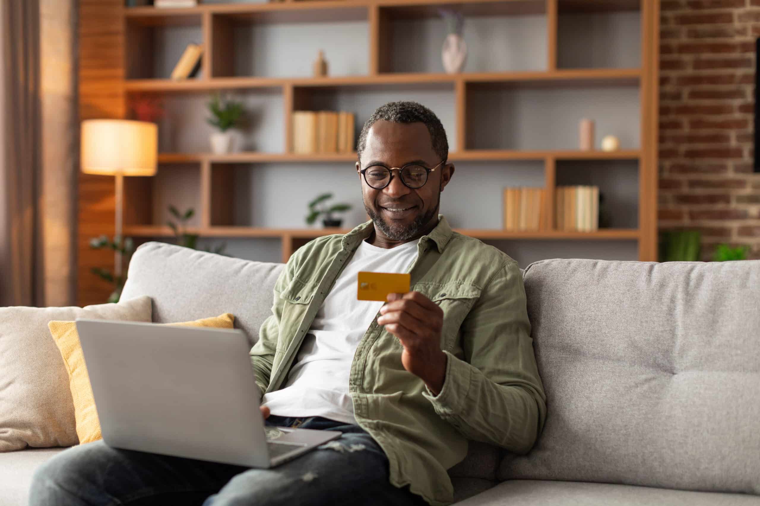 A smiling man on a couch uses a laptop and holds a credit card, likely shopping online. Bookshelves and warm lighting are behind him.