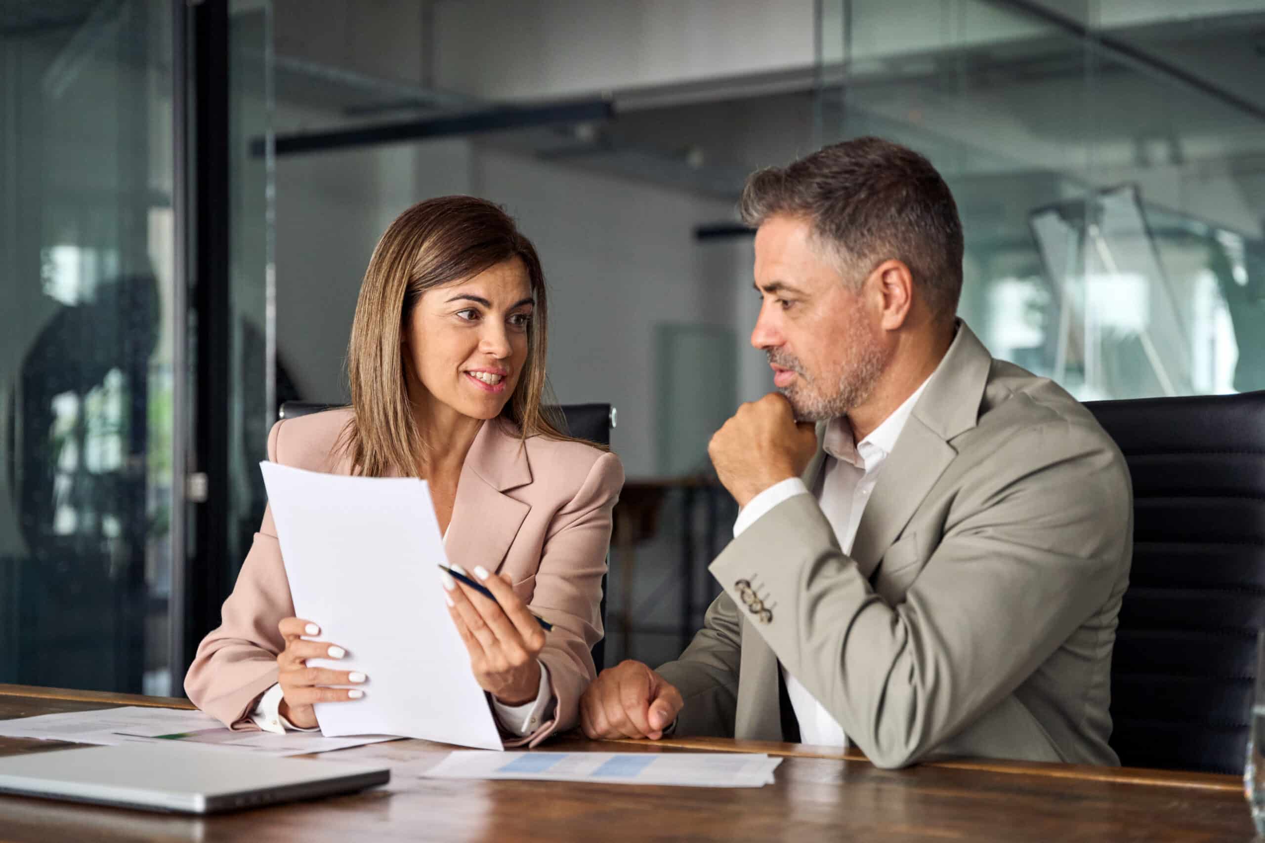 A woman and man in business attire sit at a modern office desk, reviewing documents. The woman holds papers; the man listens intently.
