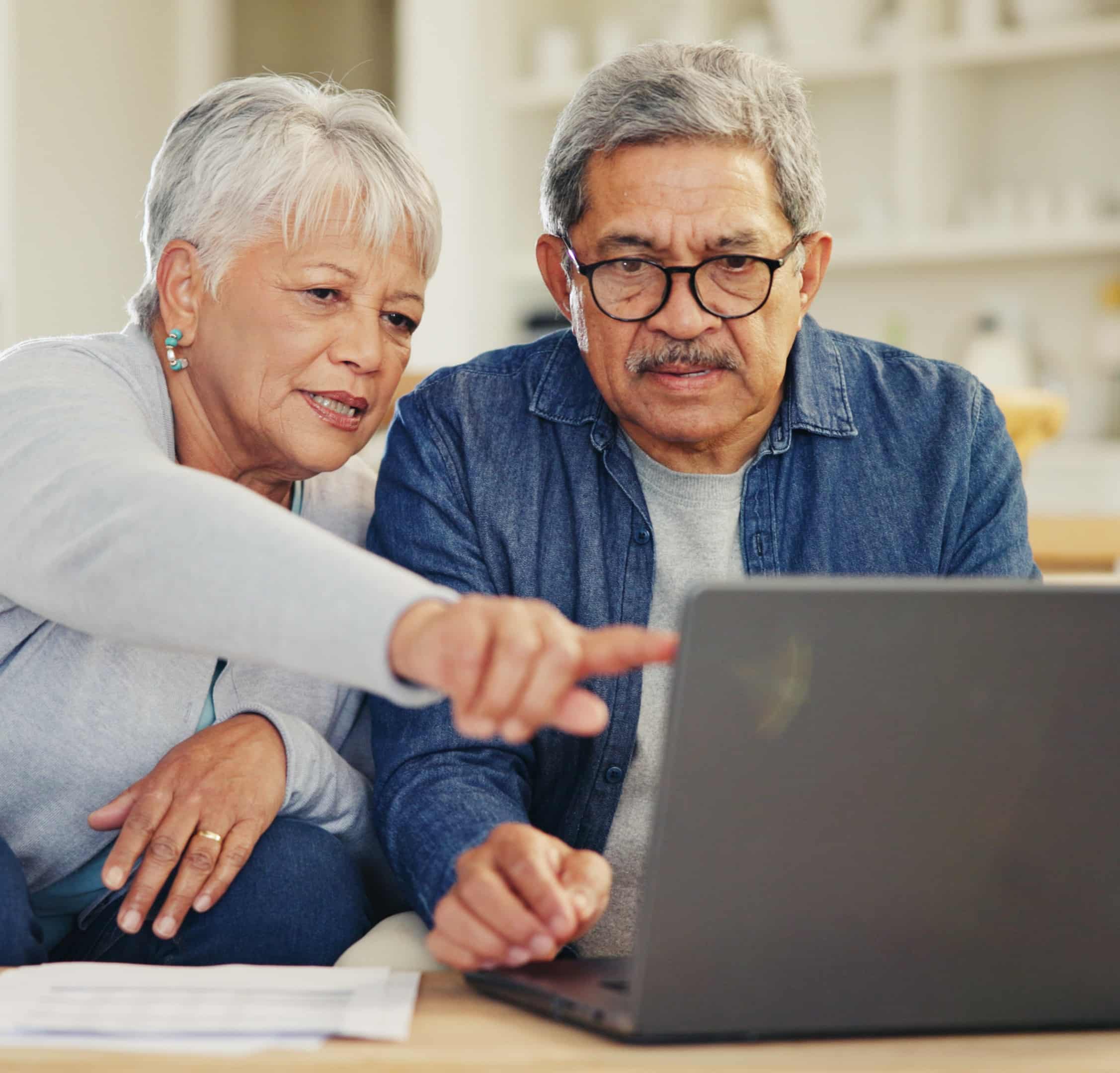 An older woman points at a laptop screen while an older man observes; both look focused. Papers lie on the table indoors.