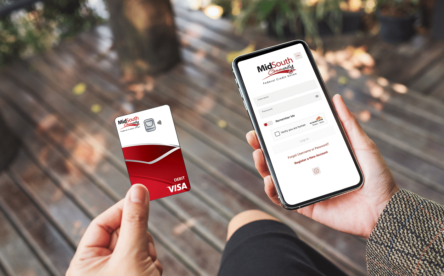 A person seated outdoors on a wooden deck holds a red MidSouth Community debit Visa card and a smartphone showing the login screen.
