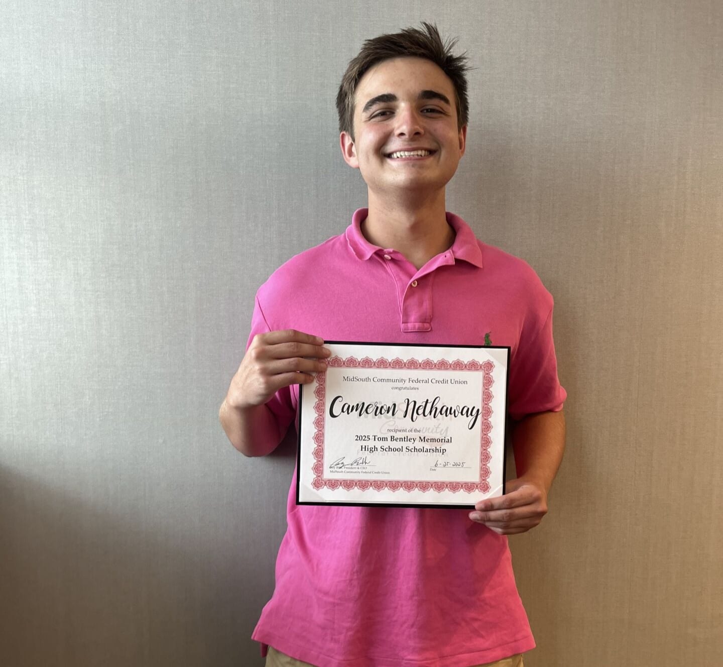 A smiling young man in a pink polo shirt holds a framed certificate for a high school scholarship by Members Community Credit Union.