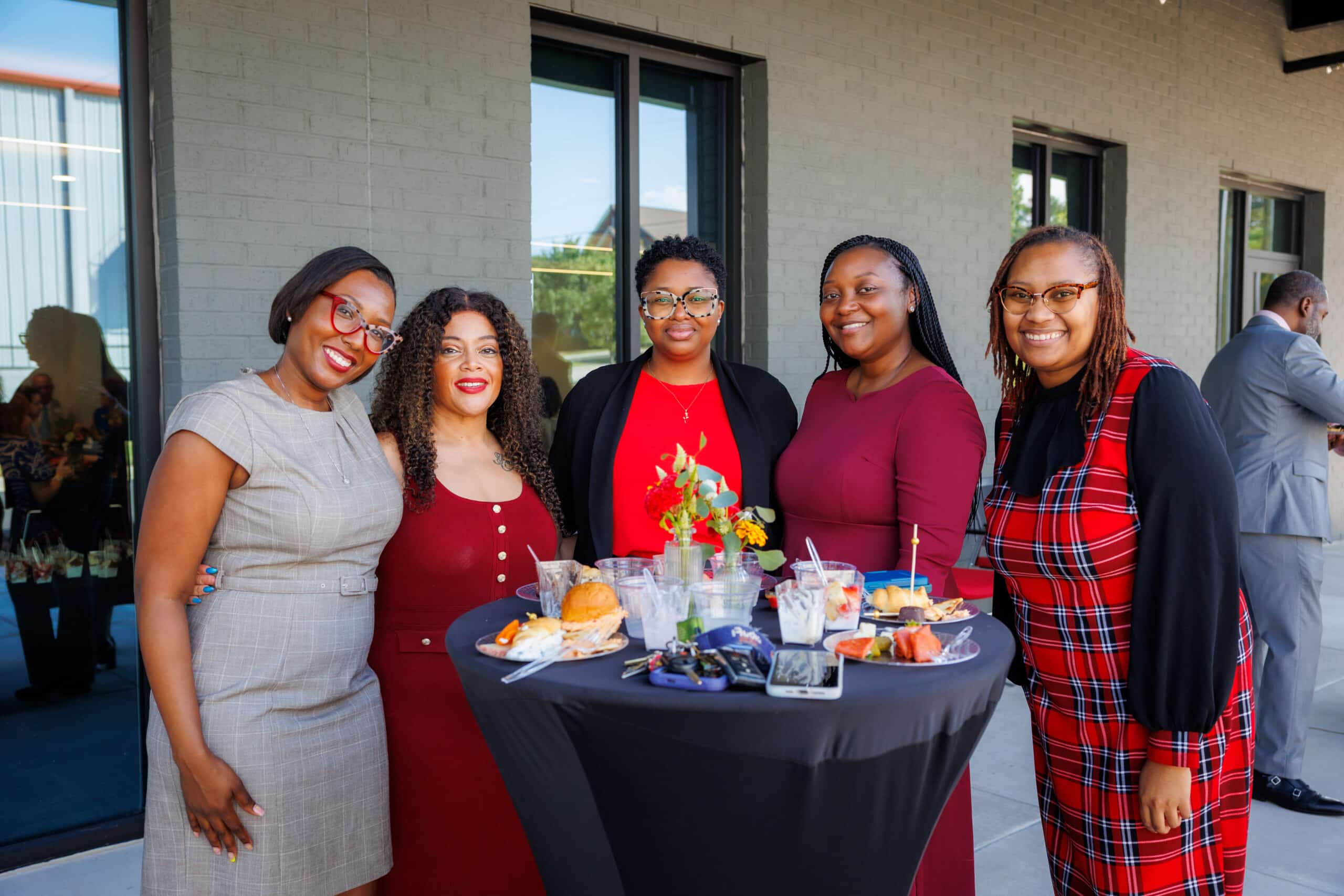 Five women smile around a high-top table with food and drinks at an outdoor event, dressed in business or casual attire.