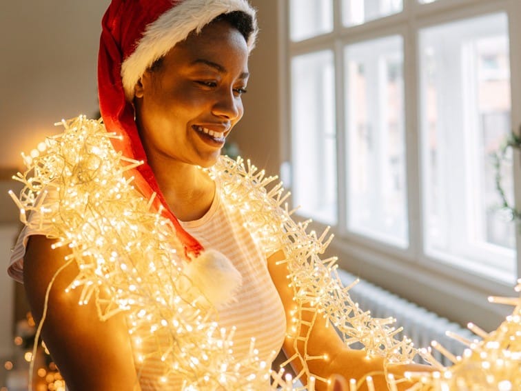 A woman in a Santa hat smiles as she wraps herself with glowing Christmas lights, standing inside near a window.