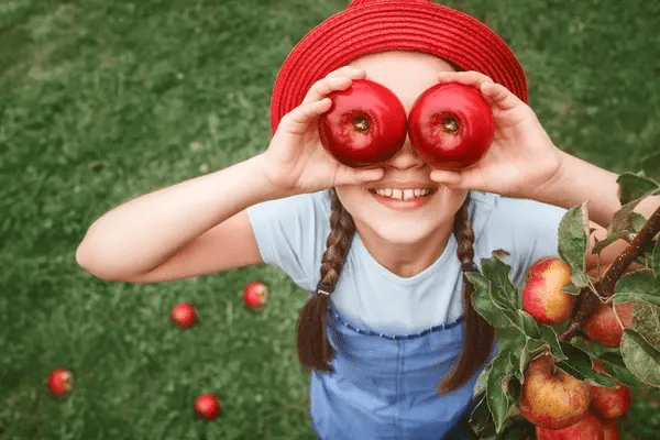 A smiling girl in a red hat playfully holds two red apples over her eyes, standing on grass with apples scattered and branch in hand.