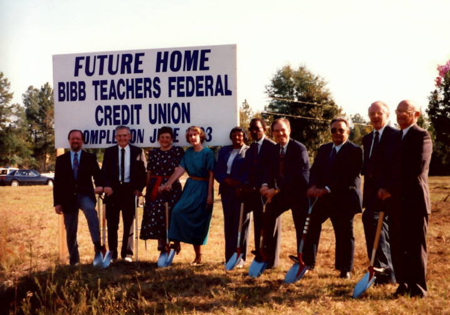 Nine people in formal attire stand in a line holding shovels before a sign for Bibb Teachers Federal Credit Union at a groundbreaking.