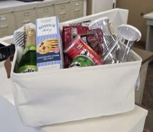 A white fabric basket with sparkling juice, cookies, tumblers, beef jerky, dog treats, and a towel on a counter near file cabinets.