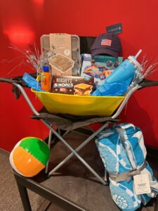Yellow folding chair displays summer gifts: s&acirc;mores kits, hat, tumbler, sunscreen, snacks, pool float, tinsel. Beach ball and towel in front. Red wall background.
