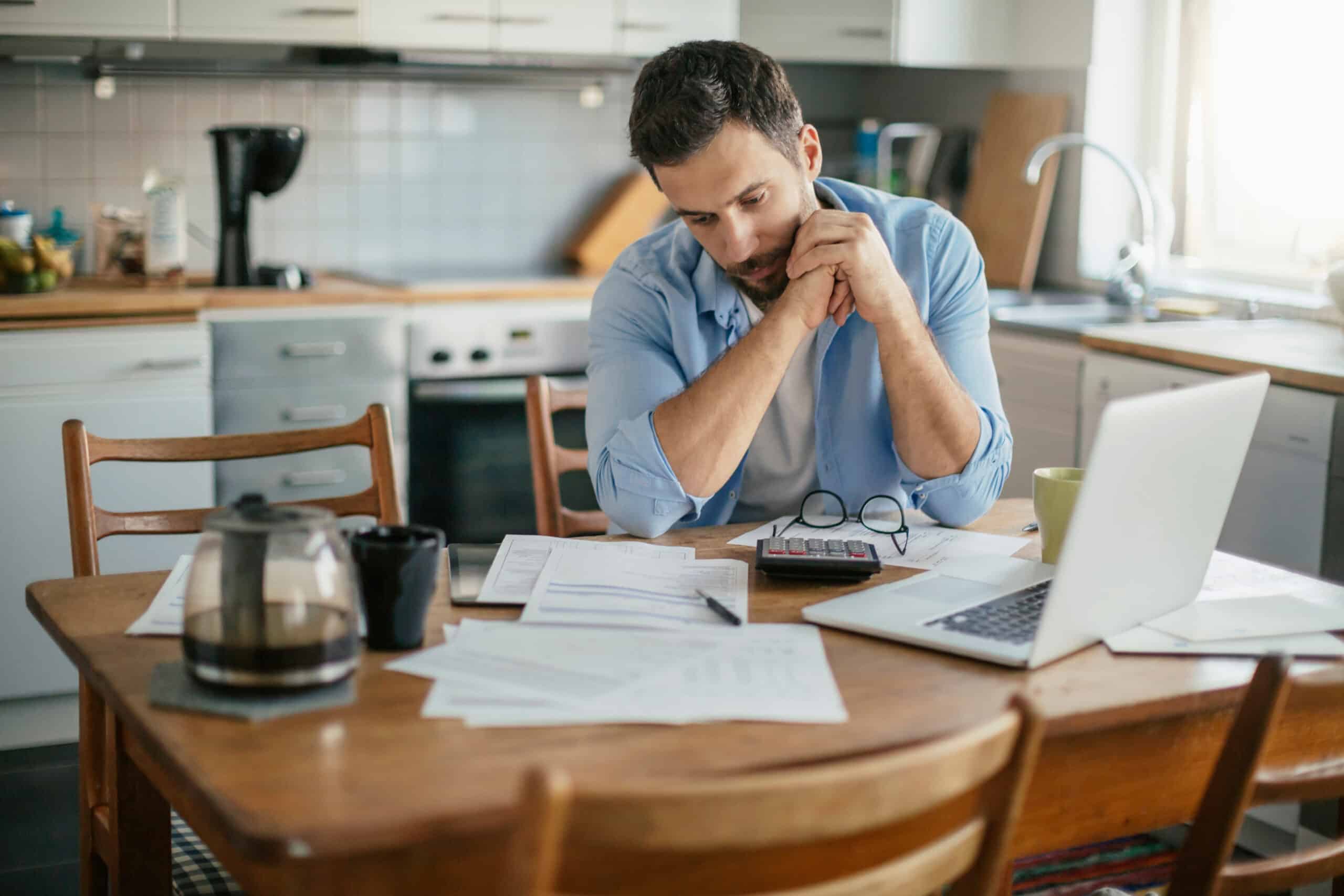 A man sits at a kitchen table cluttered with papers, calculator, glasses, coffee pot, and laptop, looking concerned reviewing documents.