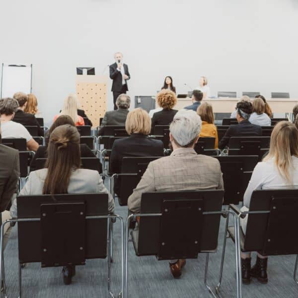 An audience faces a stage where a man speaks at a podium, with two women beside him at a table in a modern conference room.