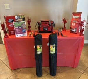 A red table holds grilling supplies like a charcoal grill kit, red cooler bag, folded chairs, utensils, charcoal bags, and decor.
