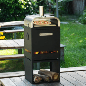A black outdoor pizza oven with a stainless top sits on a wooden patio baking pizza; firewood and green grass are in the background.