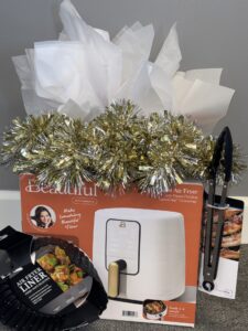 A white air fryer in a box with gold and silver tinsel and tissue paper sits by black tongs and air fryer liners against a gray wall.