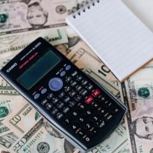 A black scientific calculator, a blank notepad, and several US dollar bills are laid out on a flat surface.