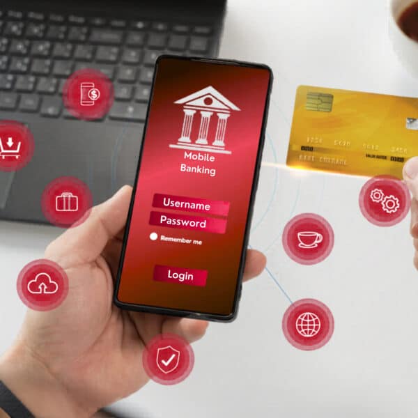 Someone holds a smartphone with a banking login screen as another hand holds a credit card, finance icons and laptop in the background.
