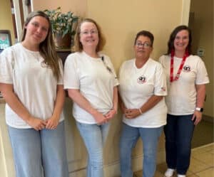 Four women are indoors, dressed in matching white T-shirts with logos and blue jeans. Three of the women wear glasses, and one sports a red lanyard. They stand smiling together in front of a beige wall and a potted plant.