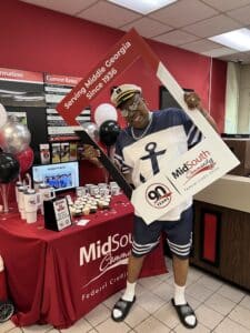 A smiling person in a sailor outfit poses with a photo frame prop at a festive table for MidSouth Community FCU’s 90th anniversary.