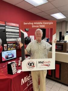 A man stands inside a bank holding a large frame with Serving Middle Georgia Since 1936 and MidSouth Community Federal Credit Union, 90 Years. The table behind him displays promotional materials and balloons.