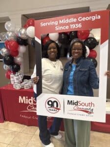 Two women smile while posing with a large photo frame celebrating 90 Years of MidSouth Community Federal Credit Union; behind them is a table with mugs, branded items, and red, black, and white balloons.