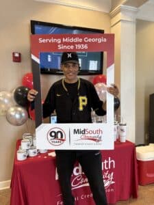 A smiling man in a black P baseball jersey holds a large photo frame prop at a credit union event with balloons and merchandise behind him.