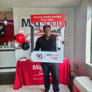 A man smiles while holding a photo frame sign celebrating 90 Years of the MidSouth Community Federal Credit Union. Behind him are balloons and a red table with the credit union’s logo and promotional items.