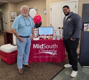 Two men stand in front of a MidSouth Community Federal Credit Union table set up with cupcakes, tumblers, and a sign celebrating 90 years of community impact.