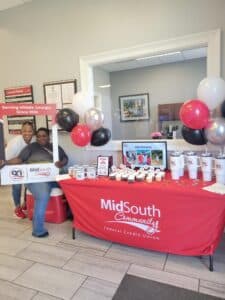 Two women pose by a MidSouth Community Federal Credit Union table with balloons, cupcakes, cups, and a display board. A large sign says Serving Middle Georgia Since 1936. The setting appears to be a bank lobby.