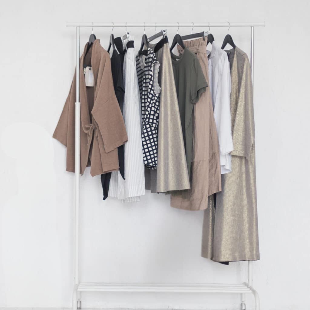 A white clothing rack holds a selection of neutral and earth-toned garments such as coats, shirts, dresses, and pants, all hanging on black hangers in front of a plain white background.